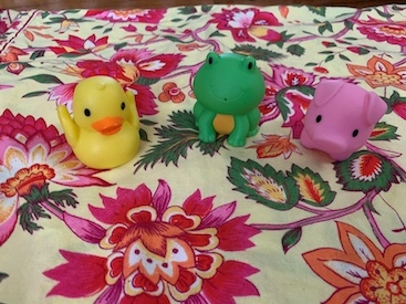 The image depicts vibrant scene featuring a toy duck, frog and pig set upon a colorful, patterned flowered cloth. 