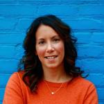 Blue brick wall in the background. A woman with dark, shoulder length brown hair is smiling and wearing an orange sweater.