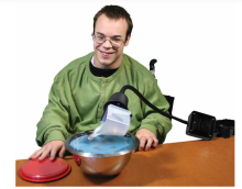 guy in wheelchair by table using the pouring cup on a flex mount to pour liquid into a bowl. 